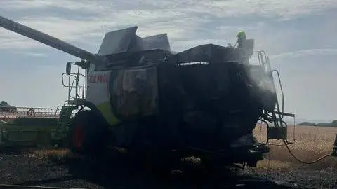 A combine harvester which has been burned in a fire sits in a large field on a sunny day. The harvested is black and there is a water hose leading up to the machine. The hay which is surrounding the machine is scorched black from a fire. 