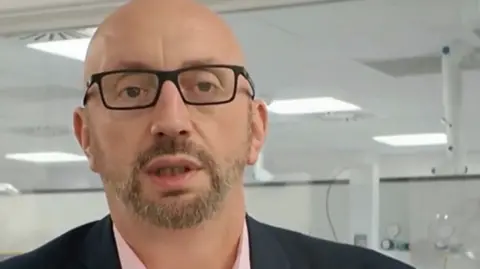 A close up image of a bald white man with a beard and black-rimmed glasses looking at a camera.