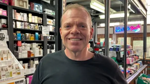 BBC Christopher Charley standing in front of his stall in a market in Coventry city centre.