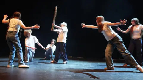 Six young actors on stage, wearing white tops and black jeans, photographed mid-way through a play