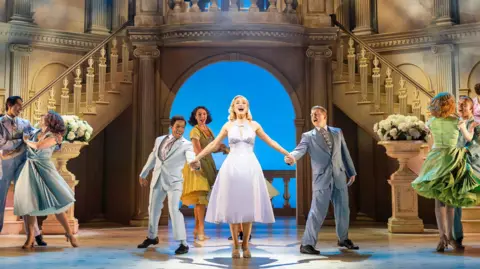 A production image of Helen George on stage, wearing a white dress with her hands being held by cast members as she sings