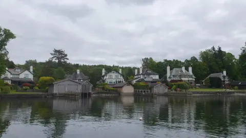 Windermere Villas comprise several large houses on the shores of the lake, some with boathouses. They are pictured from the still, dark water. A mature area of thick trees sits behind the six houses which are detached with a decent gap between each. In the main they are two-storey buildings with additional dormers and have extremely neat gardens dominated by very well-maintained hedges. The wooden, shed-like boathouses, four of which are shown, face out on to the lake and resemble double door garages with pitched roofs.