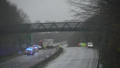 BBC The scene of the collision on the A30. There is a highways maintenance van which has its rear end destroyed. A highways maintenance small lorry is near it and there is a HGV which reads 'Conway Bailey Transport' on the side. There is a stationary police car with flashing lights behind that lorry. Debris covers the carriageway. A road sign reads Bodmin A30 straight on and left turning to Launceston, Bude, Callington and Liskeard.