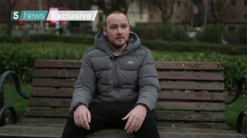 PA Media Dean Townsend has short, recending brown hair. He sits on a park bench while wearing a grey puffa jacket.