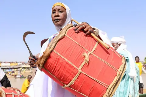 Anadolu via Getty Images A woman, in a white garment and head covering, cradles a large red drum and holds a sickle-like beater in her right hand.