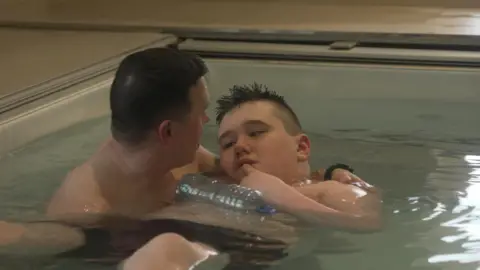 Seamus Flannigan and his son Eoin together in the swimming pool. The man has his arms around the boy as he swims in the water. 