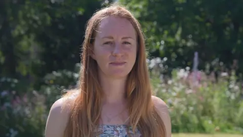 Dr Vikki Thompson looking straight at the camera. She has light brown hair. She is standing in front of a blurred background of wildflowers. 