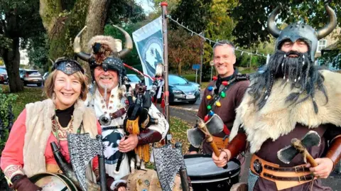 Supplied Four of the Biking Vikings stand in a line in a grassy area and smile brightly at the camera. They are wearing various Viking outfits including helmets with horns, furs and accessories with patterns on them. They are also holding their instruments. Including the pipes and drums. One Viking at the front is holding two small axes.