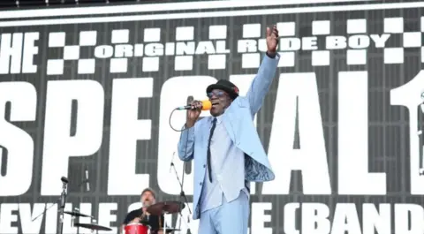 Getty Images A man wearing a blue suit and shirt stands with his arm in the air. He is holding a yellow microphone and a drummer can be seen behind him.