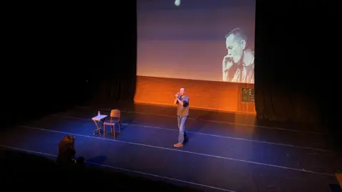 A man acting on a stage. He wears blue jeans and a grey top and is gesturing with his hands. There is a woman sitting in the front row, watching him. There is a photo of the man projected on a large screen behind.