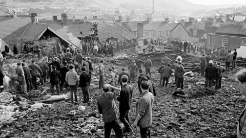 Mirrorpix via Getty Images The Aberfan disaster - crowds of men look on in this black and white photograph standing on the rubble with a digger visible in the background