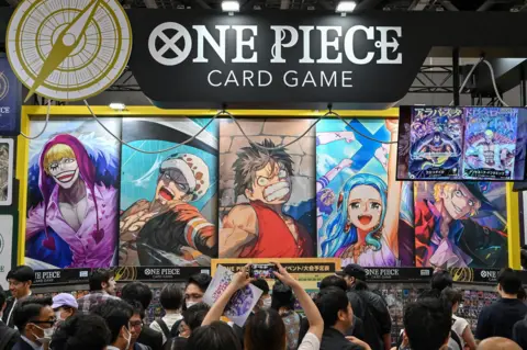 AFP via Getty Images A crowd gathers in from of a giant One Piece card display at a convention in Japan. The cards have various characters drawn in the "manga" style. A banner sits above them with the One Piece logo.