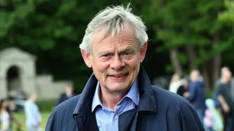 PA Media This is a photo of Martin Clunes standing in a field. He is wearing a blue suit shirt and a navy coat. 