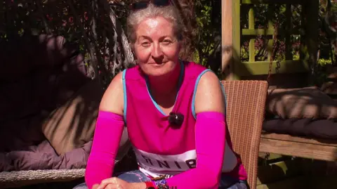 A woman wearing a pink top is sitting on a chair in a garden. She is looking towards the camera. The sun is shining on her face.