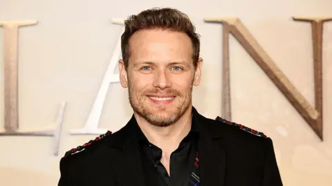Getty Images Sam Heughan with short brown hair wearing a black jacket, shirt and red and green tartan waistcoat smiling at the camera.