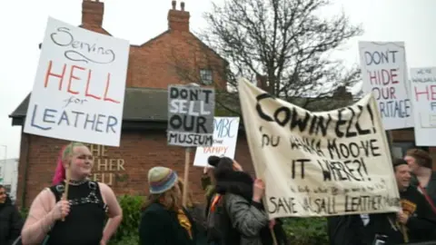 BBC A group of protestors stand outside the Walsall Leather Museum, a redbrick building with a tree and bushes outside. The protestors carry various campaign signs and banners, including 'Don't Sell Our Museum' and 'Serving Hell for Leather'