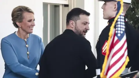 EPA Ukrainian President Volodymyr Zelensky, dressed in black, leaves the White House as a woman shakes his hand and a member of the military stands nearby with a US flag in the foreground of the image