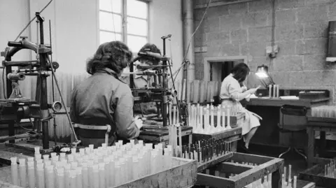 Historic England Black and white photo of two women working a lab surrounded by lots of glass bottles. You see the back of a woman with black curly hair who is looking through a microscope in the foreground. In the background you see a woman in a white-coloured outfit looking intently at an object under a bright light. 