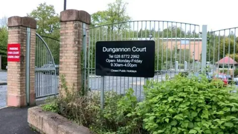 Railings outside Dungannon court car park show a black sign with the courts opening hours. Parked cars can be seen through the metal bars