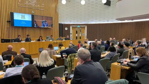 Several rows of people sat in council chamber opposite a podium