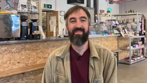 A man with short brown and grey hair and a beard and wearing a burgundy top and light brown corduroy jacket is smiling while sitting in a cafe. 