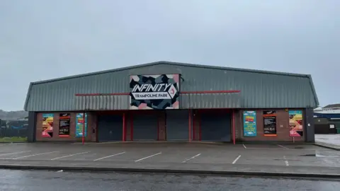 The picture show the building occupied by Infinity Trampoline Park. It is a large shed type building with a metal roof and a large sign above the front door. The car park is empty. It has been raining and the ground is wet and there are some puddles. 