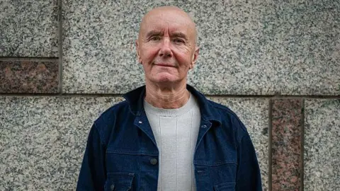 Getty Images Irvine Welsh, a bald man in a blue denim jacket and light grey jumper, smiles in front of a marble wall.