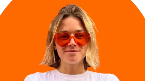 enough A woman with short blonde hair looks at the camera, wearing red love heart sunglasses. Behind her is an orange background