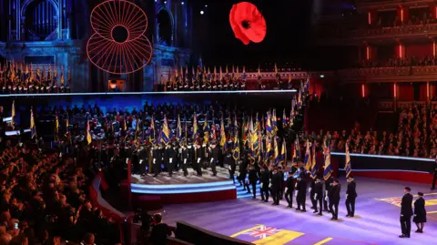 PA Media flag bearers carry flags at the Royal Albert Hall during last year's Festival of Remembrance, with two large poppies above. Hundreds of people watch in the audience.