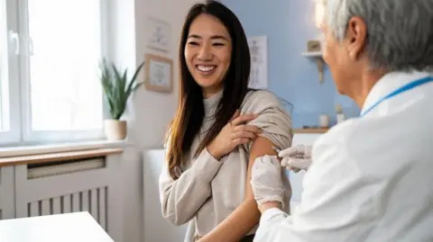 Getty Images A woman receives a flu vaccine from a doctor. The woman is holding her sleeve up and smiling and the woman giving the vaccine is wearing a white coat. The room has blue and white walls and they are sitting near a window with a plant on the windowsill.