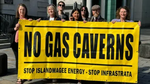 Campaigners standing outside Belfast Laganside Court complex in June 2024 holding a large yellow and black banner reading "No gas caverns" 