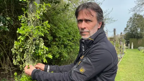 Clare Worden/BBC Alan Power stands next to a wooden pole with roses climbing up it. Some of them have withered and died away. Power has grey hair and is wearing a black jacket.