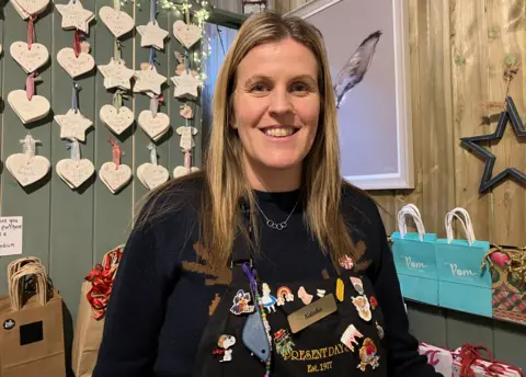 A woman with long blonde and brown hair is smiling at the camera. She is wearing a dark top and bags are behind her near the bottom of the photo.