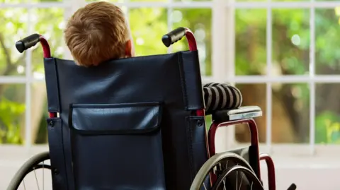 A child in a wheelchair, with his back facing the camera, looking out of a window. The chair has a black leather back and a red frame.