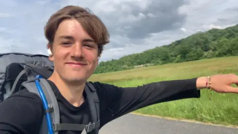 A young man skateboards along an open road lined by long grass and trees. The skateboard is not visible but he has his arms outstretched to balance himself