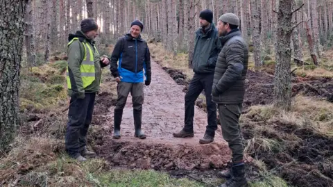 Four men standing on a new section of path running through a forest. They're wearing boots, wellies and warm clothes.