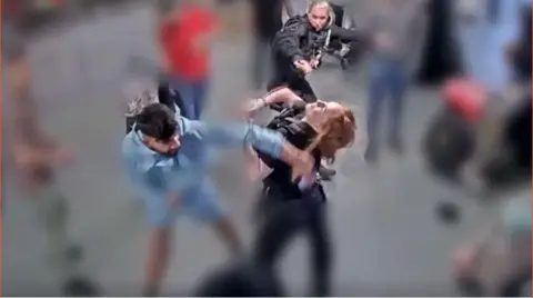 A man in blue strikes a police officer while another watches on. 