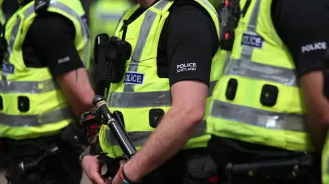 Close-up view of several Police Scotland officers wearing high-visibility yellow vests and black uniforms, standing together outdoors. One officer in the foreground holds a police baton and wears a body-worn radio, while reflective strips and police insignia are visible on the uniforms.