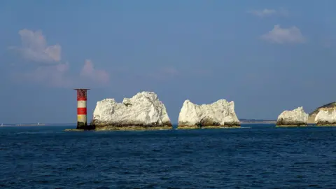 Alison Treacher A red and white stripped lighthouse with a helicopter landing pad on top is at the end of the white Needles cliff stacks. The rock formations stick out into the sea that is a bright blue. Overhead there is a light blue sky with fluffy white clouds. 