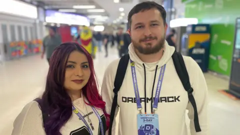 A person with long purple and pink hair wearing a black and white jersey and DreamHack lanyard stands next to a man in a white DreamHack hoodie with a three-day pass badge visible. They are standing in a wide NEC corridor with other attendees visible in the background. Both are wearing backpacks and looking directly at the camera.