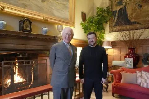 Reuters, King Charles wears a gray suit standing next to Volodimir Zellinsky in a wooden wooden sitting room with a fireplace and a red sofa with striped cushions.