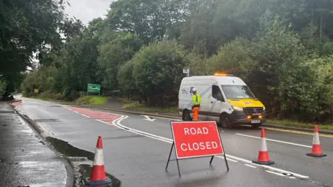 A road closed sign stands in front of a service van, with cones blocking the road amid lush greenery and overcast skies.