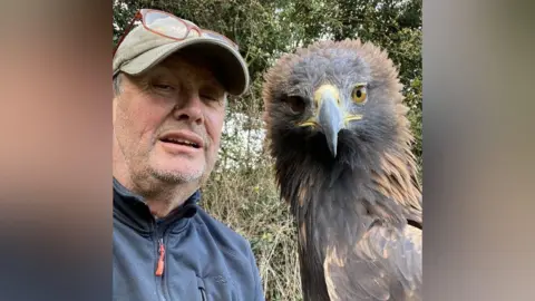Steve Cross A selfie of Steve, who is wearing a cap with glasses on top of it and a navy jumper. He is next to a large bird of prey with a yellow and black beak and brown feathers.