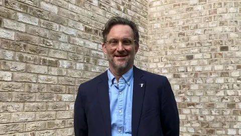 Emma Howgego/BBC Robert Pollock stares at the camera. He wears a blue shirt and a navy jacket. He stands in front of a brick wall. 