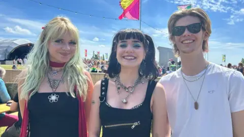 Pebble Daisy, a young woman with dark hair, lots of jewellery and a black top, poses next to her bandmates, a woman on the left with yellow and green hair and a man wearing a white t-shirt and sunglasses