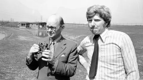 A black and white photograph of Sir Peter Scott visiting the wetlands on 20 May 1975. He is wearing a suit and tie and holding a pair of binoculars. He has glasses. Curator John Secrett is standing next to him and pursing his lips as he looks into the camera. He is a wearing a white striped shirt with a tie. The pond and the visitor centre building are behind them. 