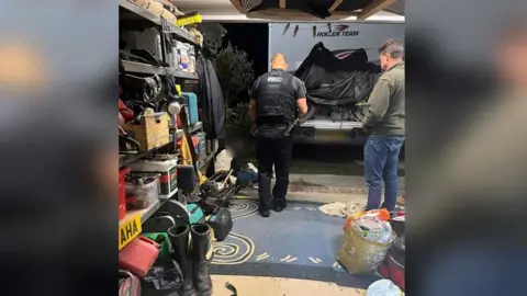 Joanne Fenton A garage with two men, one of them a police officer standing above another person whose face is blurred