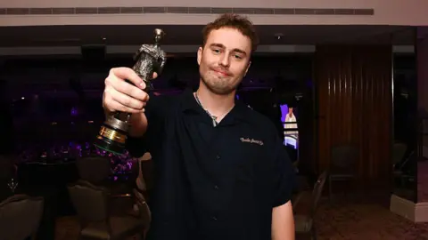 Sam Fender wears a black shirt and holds a statuette to the camera as he smiles. In the background is the ceremony hall