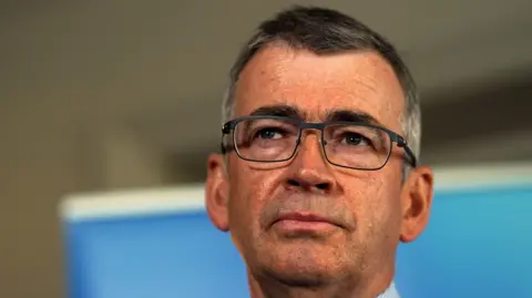 Drew Harris has short grey hair and dark framed glasses. He is wearing a white shirt. 