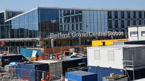 BBC Belfast Grand Central Station is a large building. The roof is zig-zag shaped. Glass windows cover the entire building. There are numerous cargo boxes and fences around the building.